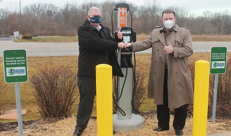 Two men in masks shaking hands in front of electric vehicle charging station